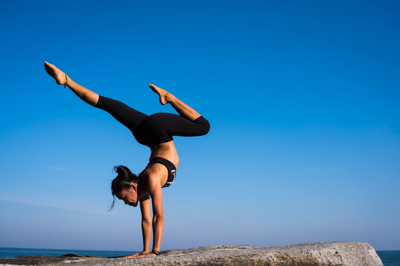 Home A woman performing a yoga handstand on a rock by the sea under a clear blue sky.